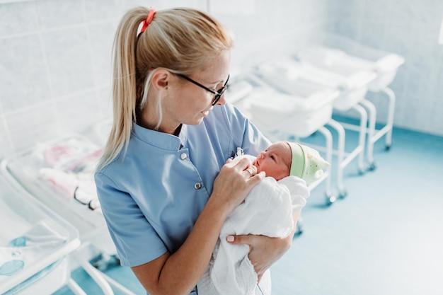 Premium Photo | Young nurse standing in maternity ward and holding newborn baby in her arms. after birth concept.